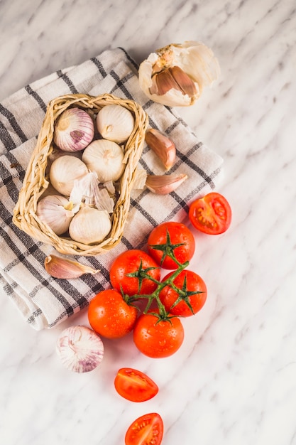 High angle view of red tomatoes; onions; garlic cloves and cloth on marble surface