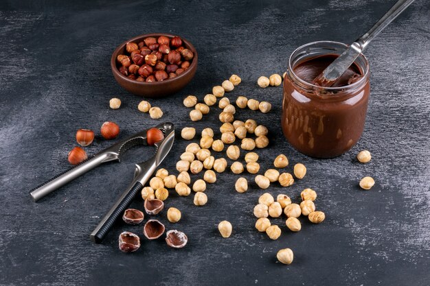 High angle view scattered cleaned hazelnuts in brown bowl with cocoa spread and nutcracker on dark stone table. horizontal