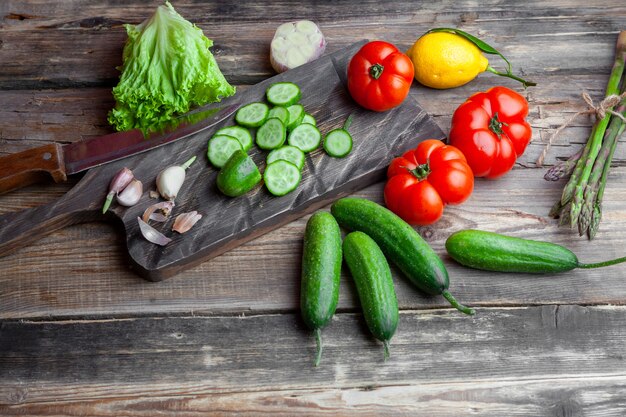 High angle view sliced cucumber in cutting board with garlic, tomatoes, lettuce, lemon and knife on dark wooden background. horizontal