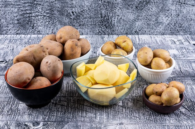 High angle view small, chopped and big potatoes in bowls on gray wooden table