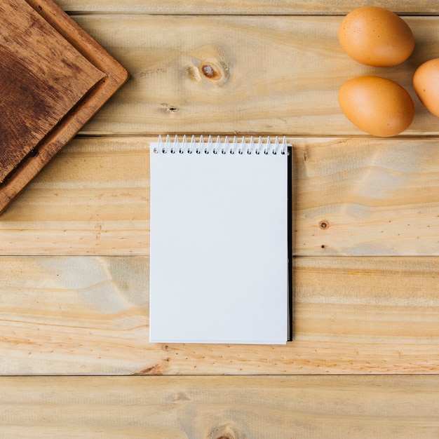 High angle view of spiral notepad; brown eggs and chopping board on wooden background