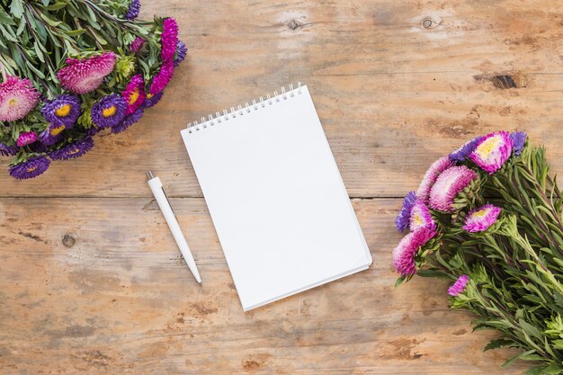 High angle view of spiral notepad; pen and bouquet of flowers on wooden desk