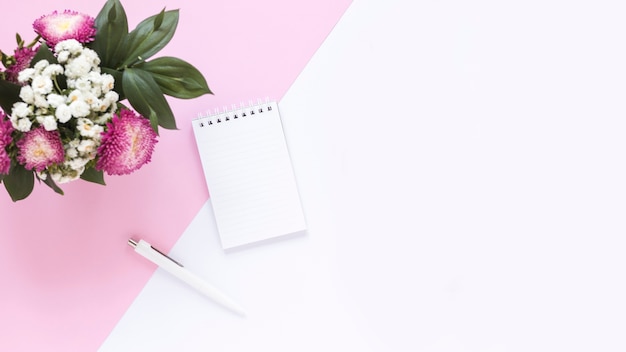 High angle view of spiral notepad; pen and bunch of flowers on dual background