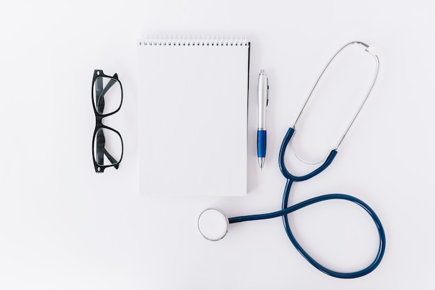 High angle view of spiral notepad; spectacles; pen and stethoscope on white surface