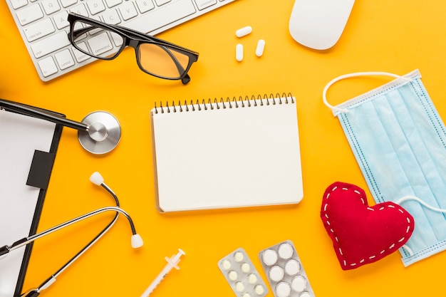 High angle view of stethoscope; injection; blister packed medicine; mask; spiral notepad; stitched heart shape against yellow backdrop