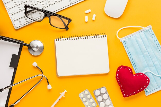 High angle view of stethoscope; injection; blister packed medicine; mask; spiral notepad; stitched heart shape against yellow backdrop