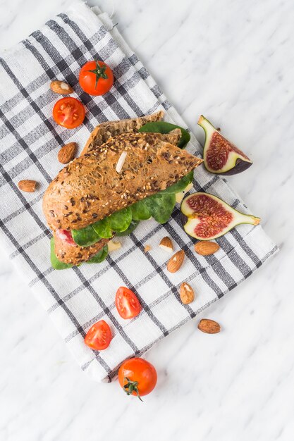 High angle view of tasty hot dog; fresh fig slices; almonds and cherry tomatoes on napkin over white backdrop