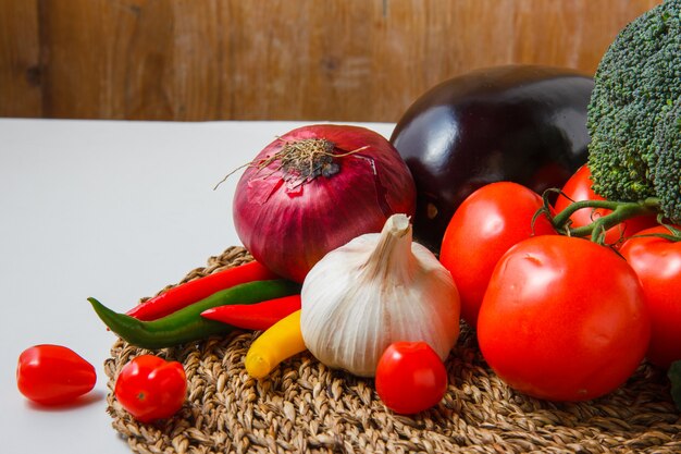 High angle view tomatoes with chili peppers, onion, eggplant, cucumber, broccoli, garlic on trivet and white surface. horizontal