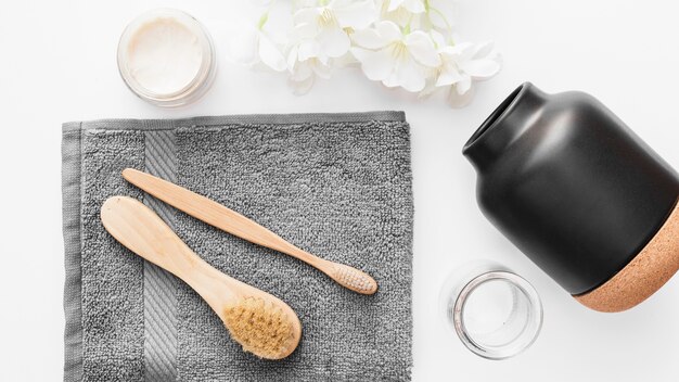 High angle view of towel; brush; moisturizing cream; flowers and jar on white background