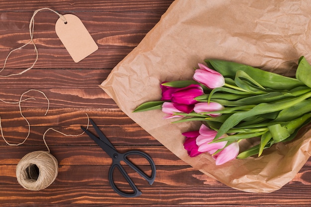High angle view of tulip flowers; brown paper; price tag; string and scissor over wooden backdrop