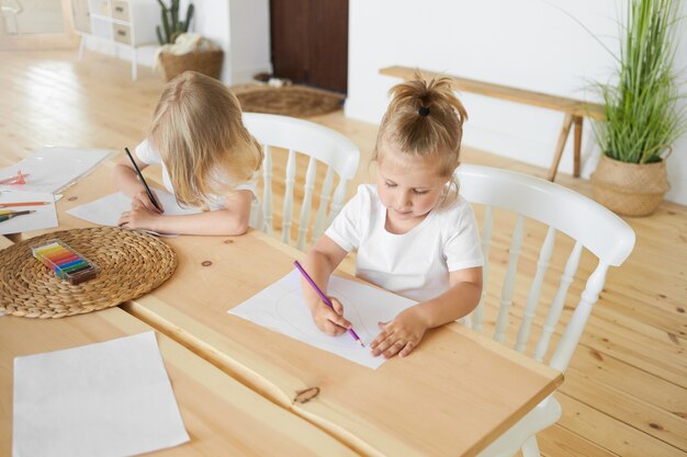 High angle view of two siblings little girl and elder brother sitting together at dining wooden table drawing images on white sheets of paper, using colorful pencils. Childhood and creativity concept