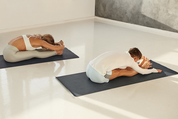 High angle view of two young muscular people male and female exercising in gym, sitting on yoga mats and bending forwards to knees, doing paschimottasana pose. Wellbeing, health and activity