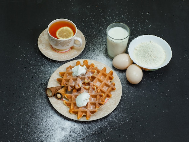 High angle view waffles in plate with tea, eggs, flour, milk on dark. horizontal