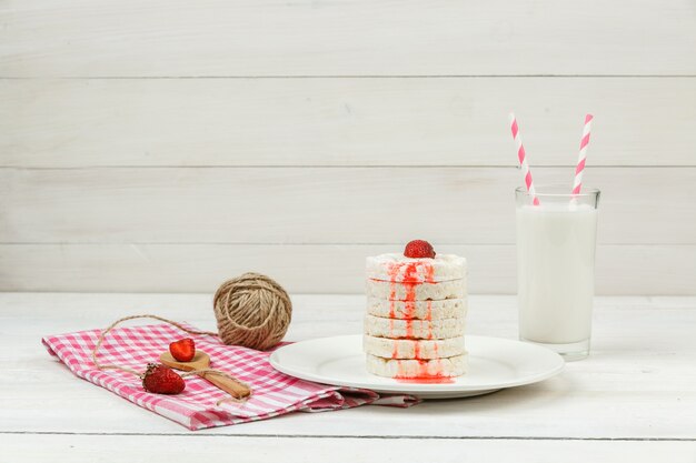 High angle view white rice wafers on plate with strawberries,meringues,clew of rope and milk on white wooden board surface.