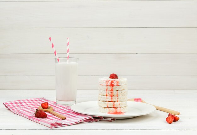 High angle view white rice wafers on plate with strawberries,wooden spoons and milk on white wooden board surface.