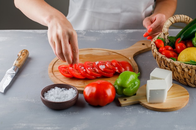 High angle view woman adding salt on sliced tomatoes on cutting board with knife, cheese, green pepper, salt on gray surface