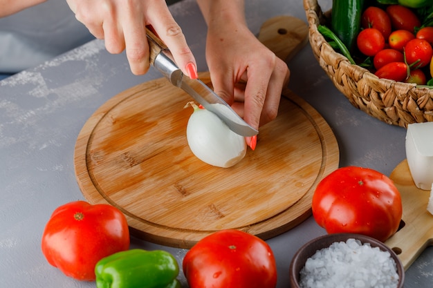 High angle view woman cutting onion into half on cutting board with knife, green pepper, cucumber, salt on gray surface
