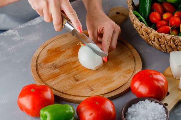 High angle view woman cutting onion into half on cutting board with knife, green pepper, cucumber, salt on gray surface