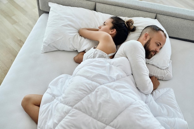 Вид под высоким углом на молодую пару, лежащую спиной к спине с проблемами в отношениях в спальне