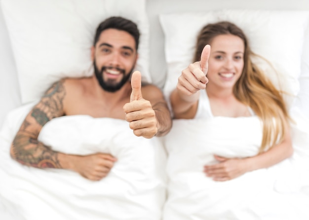 High angle view of young couple lying on bed gesturing thumbs up