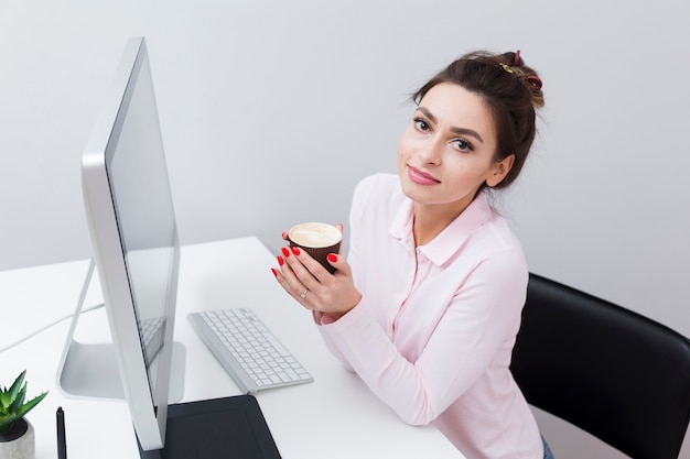 コンピューターの横にあるデスクでコーヒーカップでポーズ女性の高角