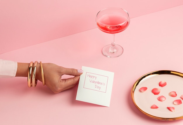 Foto gratuita alto angolo della mano della donna che tiene la carta di san valentino accanto al vetro e ai petali di rosa