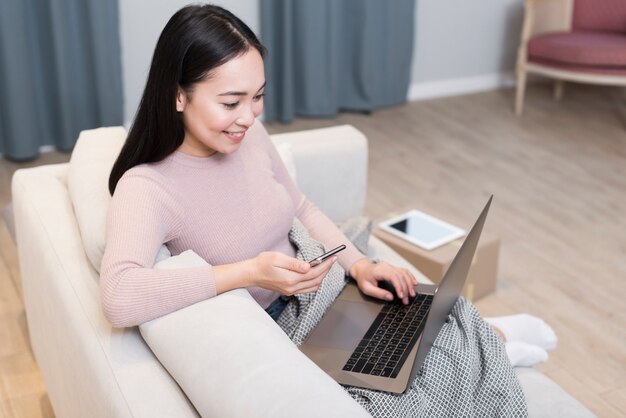 High angle of woman on the sofa holding smartphone and laptop