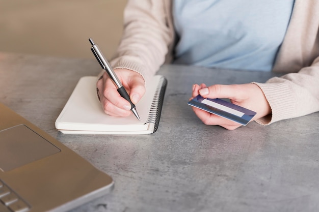 High angle of woman writing down on notebook while holding credit card