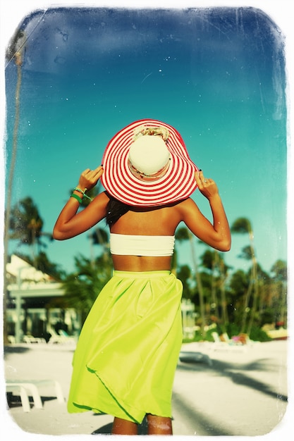 High fashion look. back of glamor sexy model girl  in retro style with palms  in colorful cloth and sunhat behind blue beach  sky