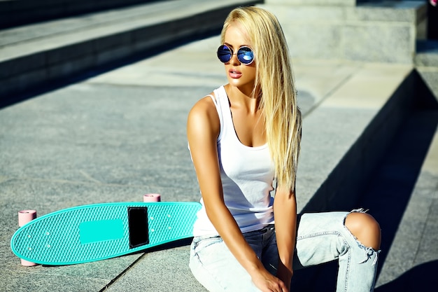 High fashion look.glamor stylish sexy beautiful young blond  model girl in summer bright casual hipster clothes with skateboard sitting in the street