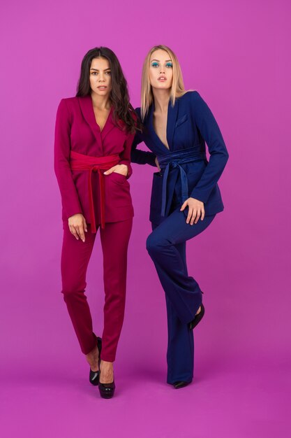 High fashion style two attractive women on violet wall in stylish colorful evening suits of purple and blue color, friends having fun together, fashion trend