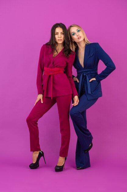 High fashion style two attractive women on violet wall in stylish colorful evening suits of purple and blue color, friends having fun together, fashion trend