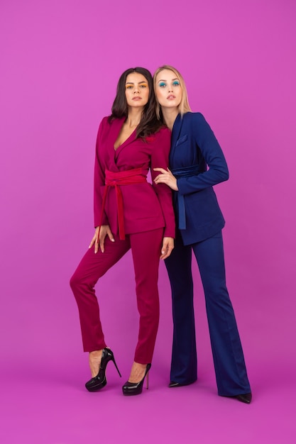 High fashion style two smiling attractive women on violet wall in stylish colorful evening suits of purple and blue color, friends having fun together, fashion trend