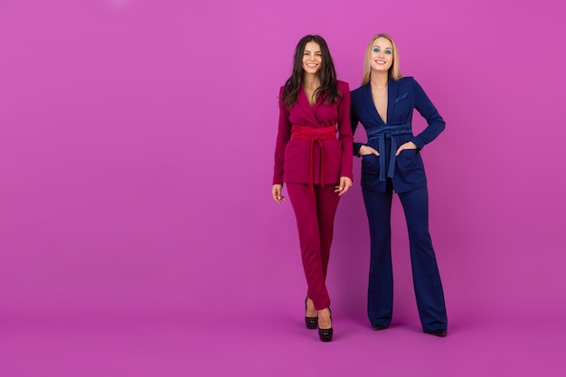 High fashion style two smiling attractive women on violet wall in stylish colorful evening suits of purple and blue color, friends having fun together, fashion trend