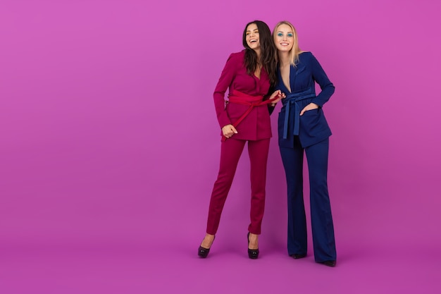 High fashion style two smiling attractive women on violet wall in stylish colorful evening suits of purple and blue color, friends having fun together, fashion trend