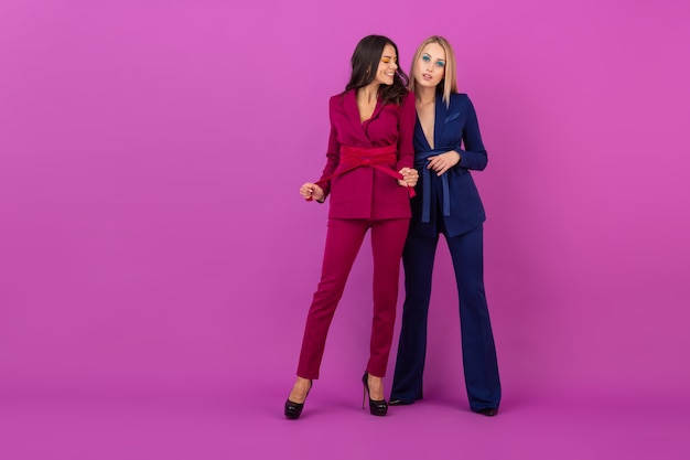 Две улыбающиеся привлекательные женщины в стиле высокой моды на фиолетовой стене в стильных красочных вечерних костюмах фиолетового и синего цвета, друзья веселятся вместе, модная тенденция