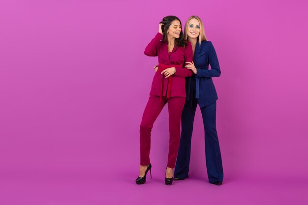 High fashion style two smiling attractive women on violet wall in stylish colorful evening suits of purple and blue color, friends having fun together, fashion trend