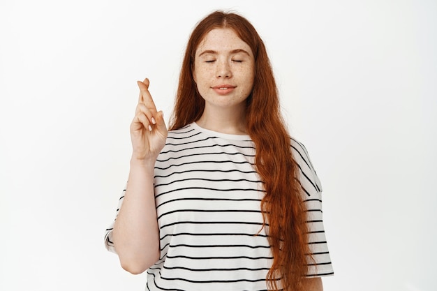 High hopes. Smiling redhead girl making wish, cross fingers and close eyes while praying, dream of smth, waiting for results, hoping to win, standing in t-shirt on white.