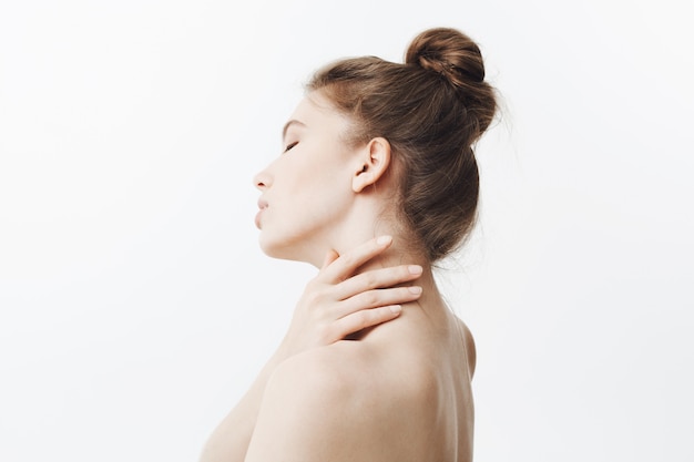 Free photo high key. stunning good-looking caucasian young woman with dark long hair in bun hairdo holding neck with hands, turning head with tired expression and closed eyes. girl suffering from pain in neck.