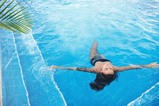 High view of sexy brunette woman lying on water in swimming pool relaxing in hotel spa near tropical trees wearing black bikini