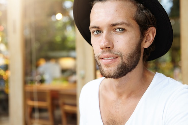 Free photo highly-detailed shot of attractive and fashionable young bearded blue-eyed male model with fair hair and clear tanned skin posing at pavement cafe wearing trendy hat