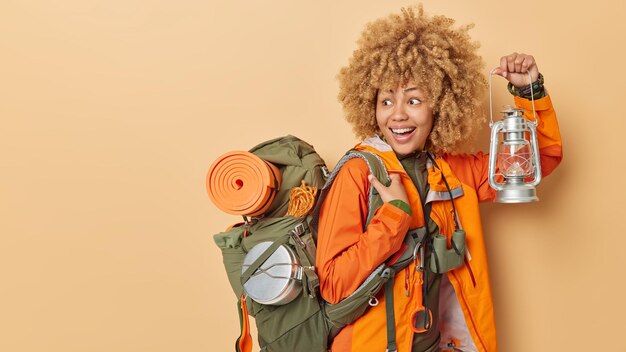 Hiking and adventure concept Positive active female camper explores new places holds lantern to see in darkness carries backpack isolated over brown background empty space for your promotion