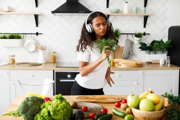 Hilarious mulatto woman with closed eyes in big headphones is smiling and emotionally pretending like she is singing in greenery near the table with fresh vegetables and fruits