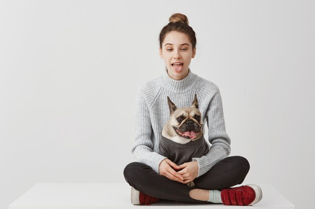 Hilarious woman 20s posing with tongue sticking out. Funny shot of young adult girl fooling around copying her dog while sitting together on table. Joy concept, copy space