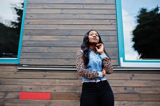 ヒョウの袖が窓のある木造家屋に対して通りでポーズをとっているジーンズシャツを着ている流行に敏感なアフリカ系アメリカ人の女の子
