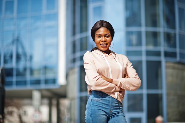 Hipster african american girl wearing pink hoodie jeans posing at street against office building with blue windows