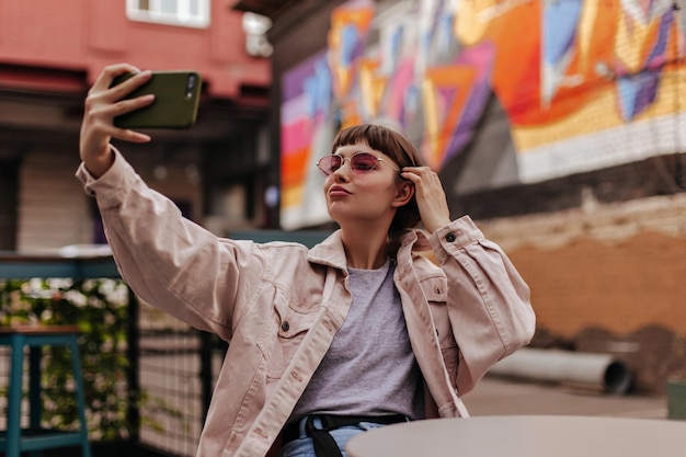 Ragazza bruna hipster che fa selfie all'aperto donna dai capelli corti in giacca beige e occhiali da sole rosa che scatta foto e si siede al bar