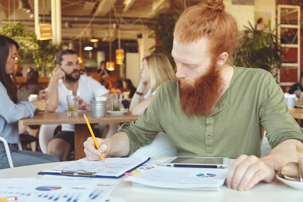 Hipster 프리랜서가 연필을 들고, 그래픽이있는 종이에 메모를 만들고, 공동 작업 공간에서 먼 곳에서 작업 할 수 있도록 디지털 태블릿을 사용합니다.