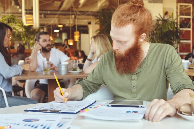 Hipster freelancer holding pencil, making notes in sheets of paper with graphics, using digital tablet for distant work at co-working space.