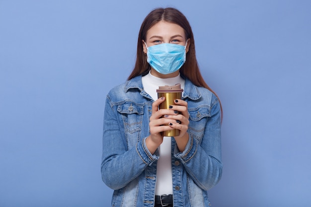 Хипстер девушка в медицинской маске и джинсовой куртке пьет кофе из термокружки